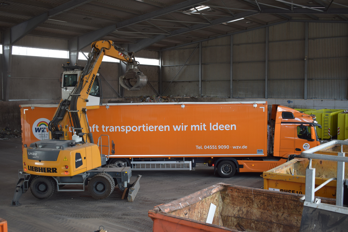 Ein großer oranger LKW wird von einem Bagger mit Müll beladen.