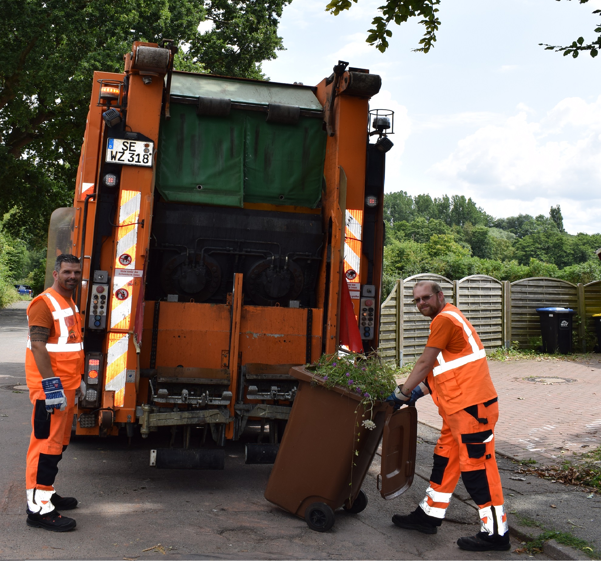Zwei Müllwerker sammeln Bioabfälle ein. Ein oranges Abfallsammelfahrzeug kippt die Tonnen.