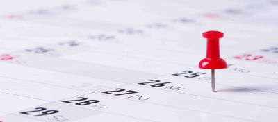 Ein perspektivisch verzerrter Kalender in Nahaufnahme zeigt in einzelnen Feldern Tage des Monats. Ein roter Steckpin markiert eines der Felder.